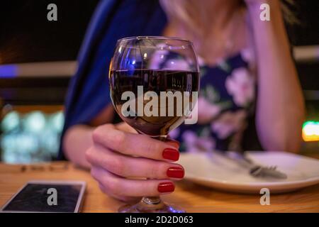 Nahaufnahme einer weiblichen Hand mit einem Glas Rotwein. Mädchen hält ein Glas Rotwein, während sie an einem Tisch in einem Café am Abend sitzt Stockfoto