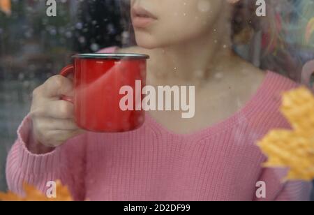 Mädchen mit roten Kaffeebecher hinter nassen Fenster mit Regentropfen Auf Glas Stockfoto