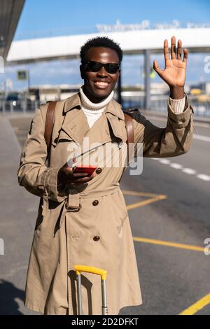 Afro-amerikanische Millennial Reisende Mann tragen beige Trenchcoat mit Koffer steht im Flughafen-Terminal, halten Handy, rufen Taxi, heben han Stockfoto