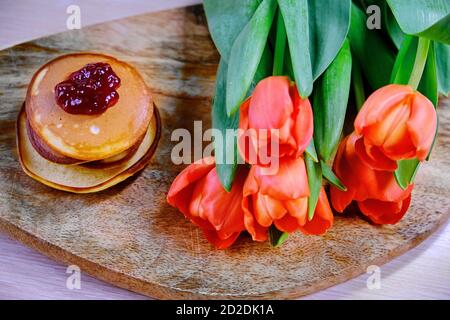 Ein Bouquet aus roten Tulpen neben einem Stapel Pfannkuchen. Heiße hausgemachte Kuchen auf Holzbrett und Blumen. Beerenmarmelade auf Pfannkuchen. Stockfoto