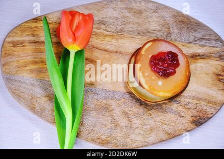 Pfannkuchen mit Marmelade und Blume. Eine rote Tulpe liegt neben dem Gebäck auf dem Küchenbrett. Ein Hügel mit frischen Pfannkuchen auf dem Tisch im Frühjahr. Stockfoto