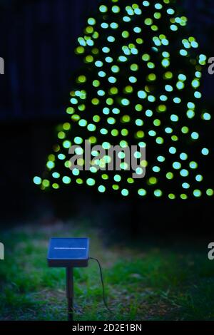 Ein Solarpanel vor einem Weihnachtsbaum mit solarbetriebenen weihnachtslichtern in der Dämmerung. Nachhaltiges Energie- und Wohnkonzept Stockfoto