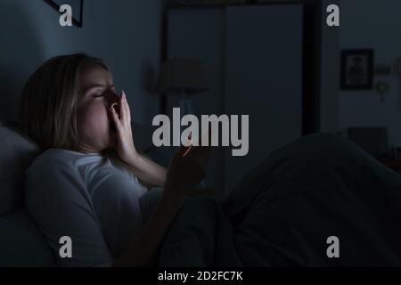 Junge schläfrige müde Frau im Bett liegend, mit Smartphone, den Mund mit der Hand und Gähnen, Augen geschlossen, kann nicht schlafen / Schlaflosigkeit, Nomophobie, Schlaf Stockfoto