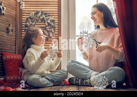 Frohe Weihnachten und schöne Feiertage! Glückliche liebevolle Familie sitzt am Fenster und macht Papier Schneeflocken für Dekoration Fenster. Mutter und Kind Kre Stockfoto