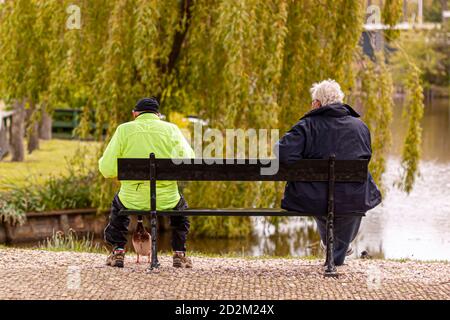 Ein junger Mann mit reflektierendem Outfit und Wintermütze und Ein älterer Mann mit grauen Haaren und Wintermantel sind Sitzen auf einer Holzbank am Avon Fluss in bat Stockfoto