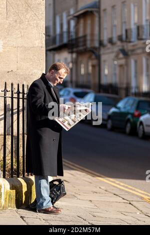 Bath, UK, 03/06/2010: Ein Kaukasier mittleren Alters, der einen langen Wintermantel trägt, steht morgens an einer Ecke und lehnt sich an Metallgeländer. Er ist c Stockfoto