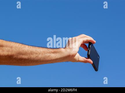 Ein abstraktes Foto, das den Unterarm und die Hand eines jungen kaukasischen erwachsenen Mannes zeigt, während er ein Smartphone hält, um ein Selfie zu machen. Es ist ein einfaches Konzept ima Stockfoto