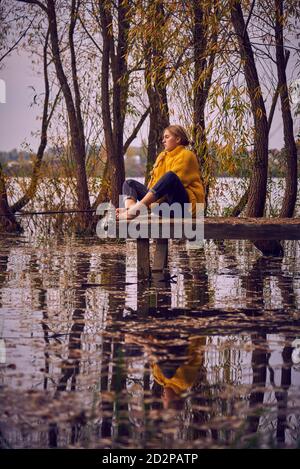 Ein junges Mädchen in einem gelben Pullover sitzt auf der Brücke über den See und fängt Fische. Stockfoto