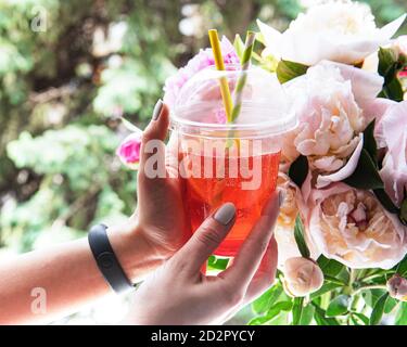 Erfrischendes Sommergetränk mit Erdbeere in weiblichen Händen vor dem Fenster und einem Bouquet von Pfingstrosen Stockfoto