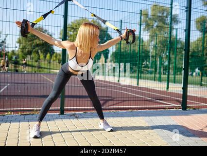 Durchführen von TRX-Übungen. Junge athletische Frau mit perfektem Körper in Sportbekleidung trainiert ihre Arme mit trx Fitnessbändern im Stadtpark Stockfoto