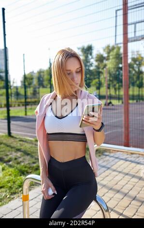 Junge athletische Frau mit perfektem Körper in Sportswear hält Handy im Stadtpark. Sportlerin, die sich nach dem Training ausruhte Stockfoto