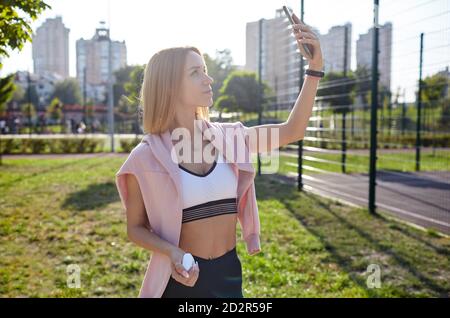 Selfie-Zeit. Junge athletische Frau mit perfektem Körper in Sportkleidung macht Selfie durch ihr Smartphone im Stadtpark. Stockfoto