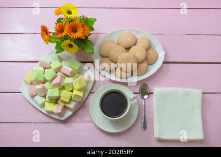 Tasse schwarzen Kaffee zum Frühstück. Marshmallows und hausgemachte Kekse auf rosafarbenem Holztisch. Romantische Tischkulisse. Stockfoto