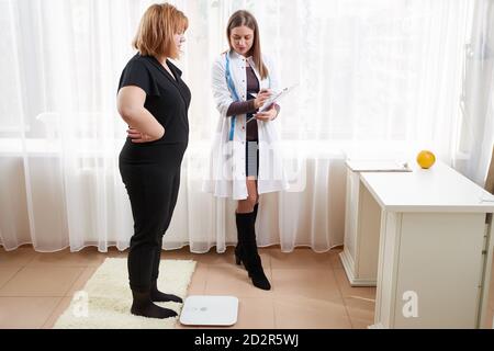 Schöne junge Ernährungsberaterin Frau Wiegen und Messen seine Patientin an Die Skala Stockfoto