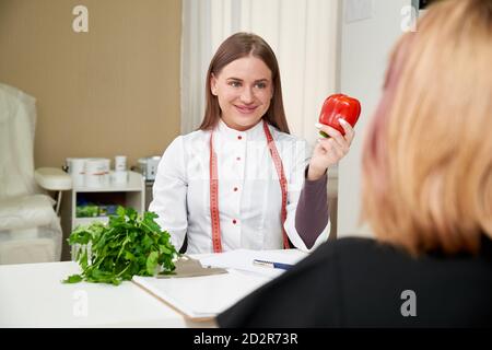 Schöne junge Ernährungsberaterin trägt weiße Uniform hält roten Paprika Stockfoto
