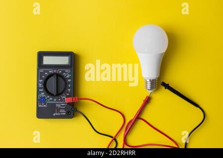 Dunkles Digitalmultimeter mit Messspitzen und LED-Lampe auf gelbem Hintergrund. Energiesparkonzept. Ein Multimeter ist ein elektronisches Messgerät Stockfoto