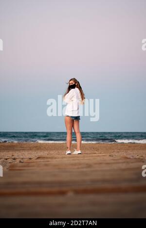 Rückansicht der weiblichen Touristen mit Schutzmaske stehen auf Sandstrand in der Nähe des Meeres gegen Sonnenuntergang Himmel und Blick über Schulter während Coronavirus-Epidemie Stockfoto