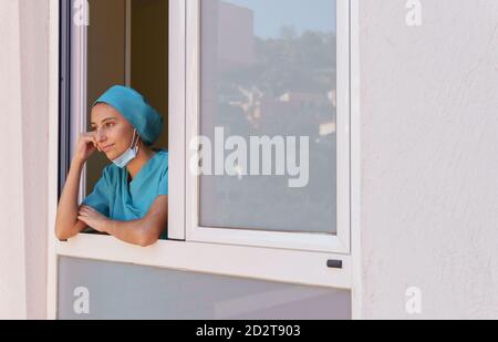 Nachdenkliche junge Ärztein Uniform mit Schutzmaske Auf dem Kinn stehend auf dem Fenster und Blick weg pensives während Sich während der Arbeit im Krankenhaus ausruhen Stockfoto