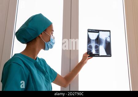 Seitenansicht der jungen medizinischen Fachärztin in OP-Kittel Und Schutzmaske Untersuchung Röntgenbild des Halses zervikal Wirbelsäule des Patienten im Krankenhaus Stockfoto