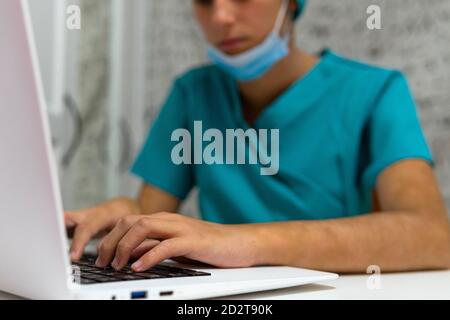 Junge Krankenschwester in blauer Uniform sitzt am Tisch und mit Laptop während der Arbeit im Krankenhaus Stockfoto