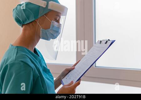 Ernsthafte weibliche Sanitäter in schützenden Gesichtsschild und Maske Lesen Zwischenablage während der Arbeit im Krankenhaus während Coronavirus Pandemie Stockfoto