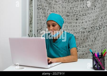 Junge Krankenschwester in blauer Uniform sitzt am Tisch und mit Laptop während der Arbeit im Krankenhaus Stockfoto