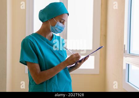 Konzentrierte junge Krankenschwester in blauer Uniform und Maske macht medizinische Hinweise zur Zwischenablage während der Arbeit im Krankenhaus Stockfoto