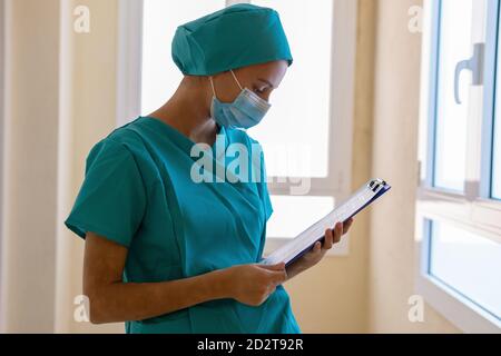 Konzentrierte junge Krankenschwester in blauer Uniform und Maske macht medizinische Hinweise zur Zwischenablage während der Arbeit im Krankenhaus Stockfoto