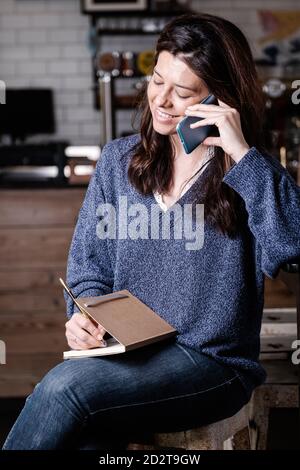 Beschäftigt weibliche Unternehmer in Freizeitkleidung sitzen mit Notebook in Cafe und sprechen über Projekt auf Handy, während Sie nach unten schauen Stockfoto