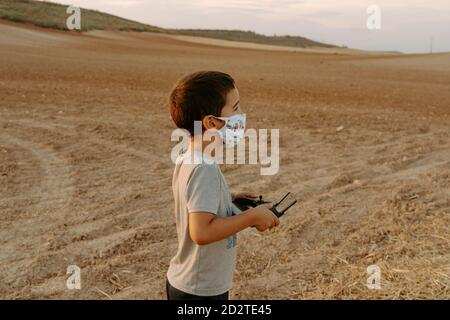Seitenansicht des fokussierten kleinen asiatischen Jungen in Schutzmaske Mit Fernbedienung während des Spielens mit fliegenden Drohne im Feld Stockfoto