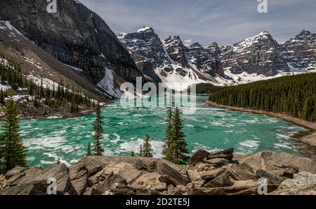 Majestätische Landschaft des gefrorenen Moraine Lake umgeben von grünen Nadelbäumen Wald und raue felsige Berge bedeckt mit Schnee in bewölkt Tag im Banff National Park in Kanada Stockfoto