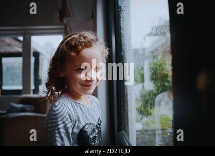 Portrait von lächelnd Schulmädchen stehen in der Nähe Fenster mit regen Tropfen auf dem Fenster in der Regenzeit Stockfoto