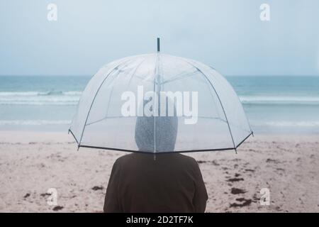 Rückansicht des nicht erkennbaren Reisenden im Regenmantel mit transparentem Schirm Wandern auf leerem Sandstrand in Richtung wehendes Meer in bewölktem Zustand Tag Stockfoto