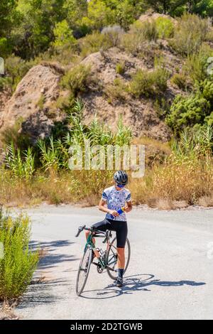 Ganzkörper-Passform weibliche Radfahrer in Sportbekleidung und Helm Frisches Wasser aus der Flasche trinken, während Sie sich nach dem Radfahren ausruhen Bergstraße hinauf Stockfoto