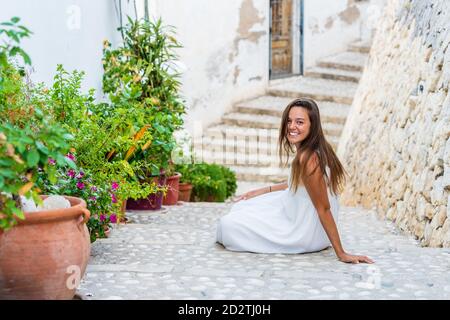 Volle Seitenansicht der entzückten jungen weiblichen Reisenden in Weißes Kleid sitzt auf einer Treppe in der Nähe Steinmauer und grün Topfpflanzen in einer engen Straße der Altstadt Stockfoto