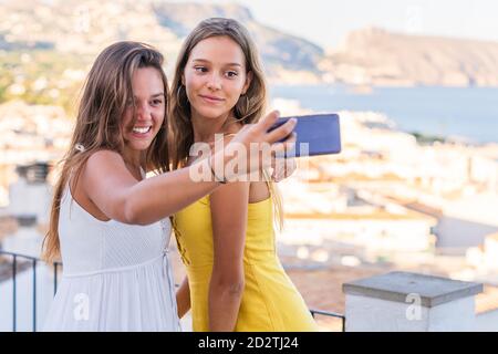 Optimistische Millennial Freundinnen umarmen und Selfie auf dem Handy nehmen Telefonieren Sie, während Sie Sommerferien zusammen in Küstenstadt in der Nähe verbringen Meer Stockfoto