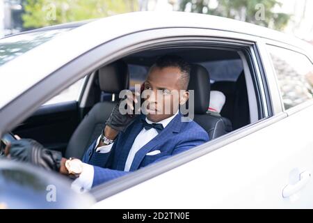 Ernst ethnischen männlichen Unternehmer in edlem Anzug mit Fliege Fahren Luxus-Auto, während auf dem Smartphone sprechen und wegschauen Stockfoto