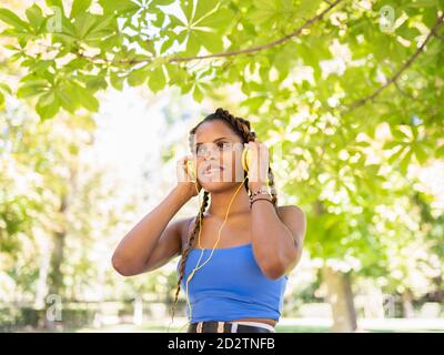 Positive hispanische Frau, die im Stehen Musik in Kopfhörern hört Im Stadtpark an sonnigen Tag und Blick weg Stockfoto