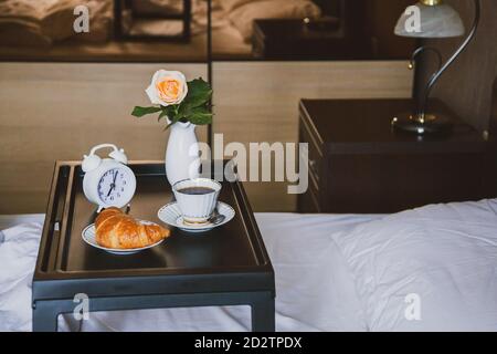 Guten Morgen. Frühstück im Bett auf einem Tablett mit Kaffee und Croissant und Rose in Vase, Holzinterieur, weißes Bett. Stockfoto