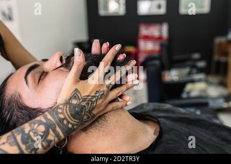 Seitenansicht des Crop anonymous tätowiert Friseur tun Massage auf Bart des entspannten männlichen Klienten während der Pflege Verfahren im Friseurladen Stockfoto