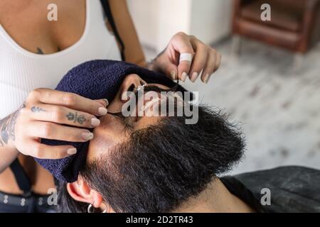 Hoher Winkel der Ernte unkenntlich weibliche Master mit tätowierten Körper Styling Schnurrbart von glücklich brutal bärtigen Mann mit Piercing während Haarschnitt Verfahren im Friseurladen Stockfoto