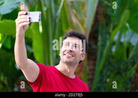 Junger Mann mit Smartphone macht ein Selfie, gekleidet mit rotem T-Shirt auf grünem Naturhintergrund am Bahnhof Stockfoto