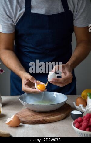 Ernte unkenntlich Mann in Schürze zerbrechen rohes Ei in Schüssel Beim Zubereiten des Frühstücks am Tisch mit Zutaten Stockfoto