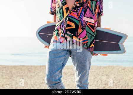 Crop-Ansicht von anonymen jungen Hipster männlichen Skater in trendy Sommer-Outfit trägt Skateboard beim Stehen auf dem Damm gegen Meer Und wolkenloser Himmel am Sommertag Stockfoto