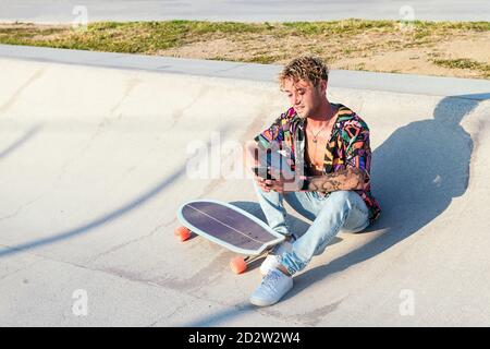 Handsome Millennial lockig behaart Kerl in unknopfed bunte Hemd und Jeans sitzen mit Skateboard auf Betonrampe und Surfen mobil Telefon während der Ruhe im Skatepark im Sommer Tag Stockfoto