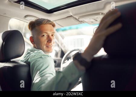 Seitenansicht eines gutaussehenden lächelnden männlichen Fahrers, der auf dem Parkplatz saß Luxus-Auto und Blick auf die Kamera Stockfoto