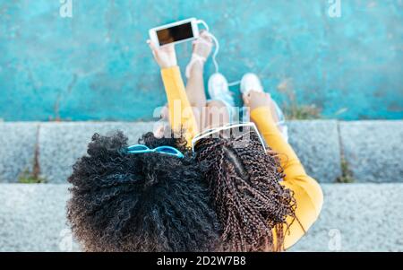 Von oben eine nicht erkennbare afroamerikanische Frau mit lockigem Haar, die auf einer Steintreppe mit einer Frau mit Zöpfen sitzt und zusammen Videos auf dem Handy ansieht Stockfoto