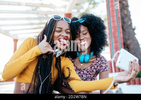 Lächelnde schwarze Frau mit Zöpfen umarmt fröhliche afroamerikanische Freundin mit lockigen Haaren und nimmt Selfie zusammen, während sie im Sommer auf der Promenade steht Stockfoto