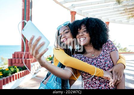 Lächelnde schwarze Frau mit Zöpfen umarmt fröhliche afroamerikanische Freundin mit lockigen Haaren und nimmt Selfie zusammen, während sie im Sommer auf der Promenade steht Stockfoto