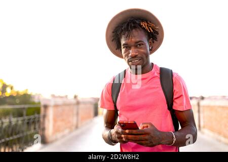 Schöner afroamerikanischer männlicher Reisender mit Rucksack, der auf der Brücke steht Und das Smartphone beim Blick auf die Kamera Stockfoto